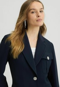 Navy blazer with structured lapels, two front pockets, and silver buttons. Model wearing earrings and a white top underneath. Smooth fabric.