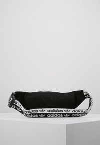 Černá látková taška zaobleného tvaru s nastavitelným popruhem. Obsahuje bílý vzorovaný popruh s logem "adidas" podél strany.