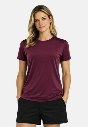 Frau mit blonden Haaren, die ein kurzärmeliges dunkelrotes Hemd und schwarze Shorts trägt, steht mit den Händen in den Taschen vor einem weißen Hintergrund.