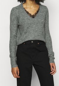 Grijze gebreide trui met een V-hals en lange mouwen, gelaagd over een zwart top met kantafwerking. Gecombineerd met zwarte high-waisted broek.