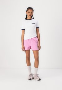 Hvit T-skjorte med marineblå detaljer og logo, kombinert med rosa shorts med snor; modell iført hvite knestøtting og lyse joggesko.