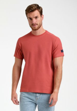 Camiseta de manga corta texturizada en color coral con cuello redondo y detalle de logotipo en el costado. Combinada con jeans de un azul claro.