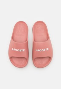 Infradito in gomma rosa con design open toe, caratterizzate da una suola texturizzata e dal prominente logo bianco "LACOSTE" sul lato.