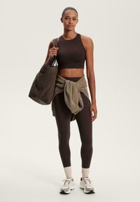 Brauner Crop-Top und Leggings, kombiniert mit einem lockeren olivgrünen Pullover, der um die Taille gebunden ist. Trägt eine große braune Tragetasche und weiße Sneaker.
