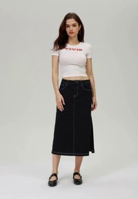 Jupe en denim noir avec des coutures blanches contrastées, de longueur genou avec une fente sur le côté ; associée à un T-shirt blanc ajusté et court.