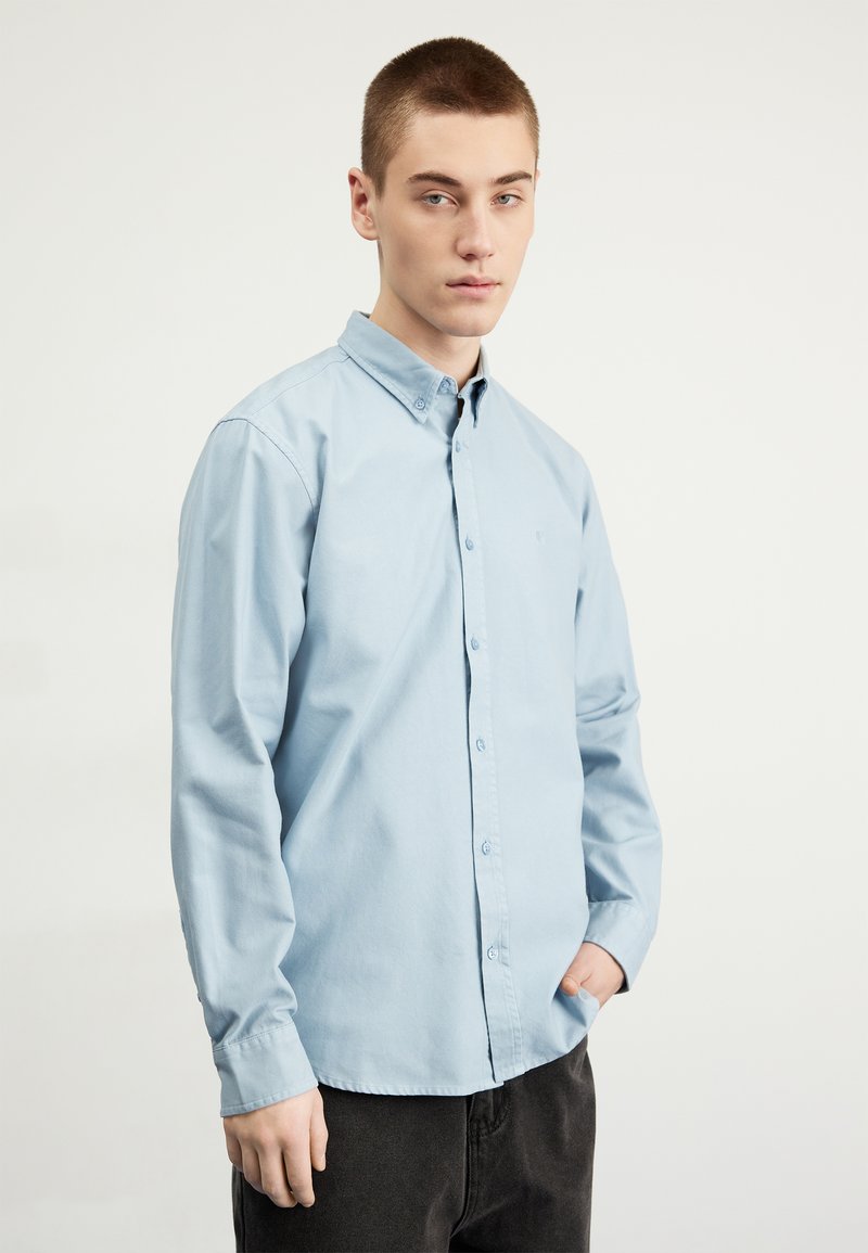 Chemise bleu clair à manches longues, avec un col et un petit logo. Le tissu semble lisse et légèrement texturé.