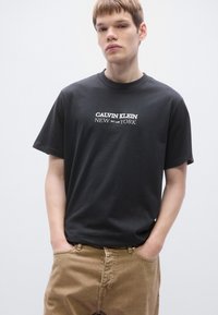 Melna kokvilnas T-krekls ar apaļu kakla izgriezumu, uz tā ir balts uzraksts: "CALVIN KLEIN NEW EST. 1968 YORK." Īsas piedurknes un brīvs piegriezums.