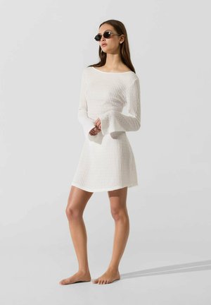 Femme portant des lunettes de soleil noires et une mini-robe blanche texturée à manches longues, debout pieds nus sur un fond clair uni.