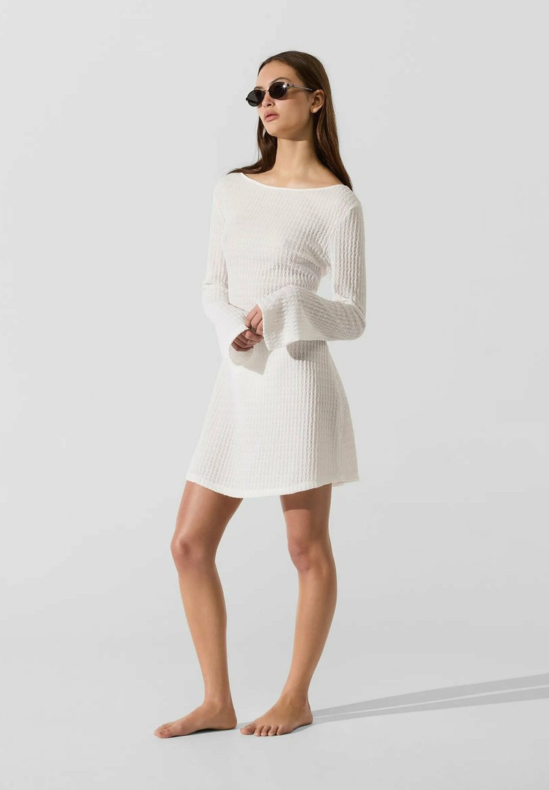 Femme portant des lunettes de soleil noires et une mini-robe blanche texturée à manches longues, debout pieds nus sur un fond clair uni.
