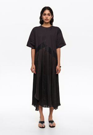 Mujer con cabello largo y oscuro que lleva un vestido negro suelto de manga corta con detalles de encaje, de pie mirando hacia adelante con sandalias negras de punta abierta.