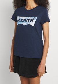 Námorně modré bavlněné tričko s bílým logem Levi's nad pozadím s obláčkovým vzorem, klasickým kulatým výstřihem a krátkými rukávy.