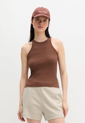 Giovane donna che indossa un top marrone senza maniche a coste, pantaloncini beige e un cappellino marrone abbinato Marc O’Polo, in piedi davanti a uno sfondo semplice.