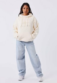 Vrouw met lang ingevlochten haar, gekleed in een off-white hoodie, lichtblauwe wijde jeans en witte sneakers, staand tegen een effen lichte achtergrond.
