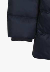 Rukáv námořnickomodré péřové bundy s hladkým, lesklým texturem a ribbovaným pleteným manžetou pro těsné přilnutí. Šikmé švy probíhají podél rukávu.