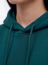 Person trägt einen dunkelgrünen Hoodie mit locker gebundenen Kordeln nahe am Hals, wobei ein Teil des unteren Gesichts und des Halses sichtbar ist.