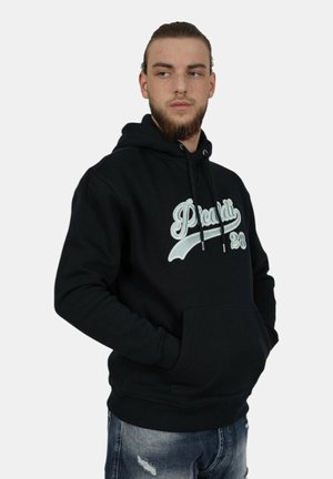 Mann mit Bart und zurückgebundenem Haar, trägt einen schwarzen Hoodie mit der Aufschrift "Picardi 28", die Hände in den Taschen und zerrissene blaue Jeans, schaut zur Seite.