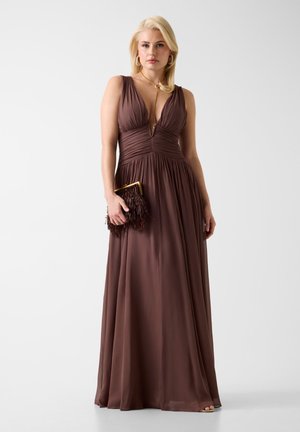 Kobieta w brązowej sukni bez rękawów z głębokim dekoltem w serek, trzymająca fakturowaną kopertówkę, nosząca kolczyki koła i długi złoty naszyjnik na jednolitym tle.