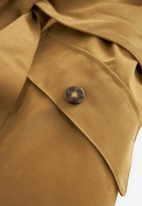 Bruine stof met een gladde textuur, voorzien van een gebogen flap en een donker, schildpadmotief knop, die subtiele stikdetails benadrukt.