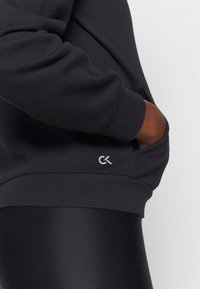 Personne portant un sweat-shirt noir Calvin Klein, main dans la poche avant et petit logo blanc CK sur le bord de la poche.