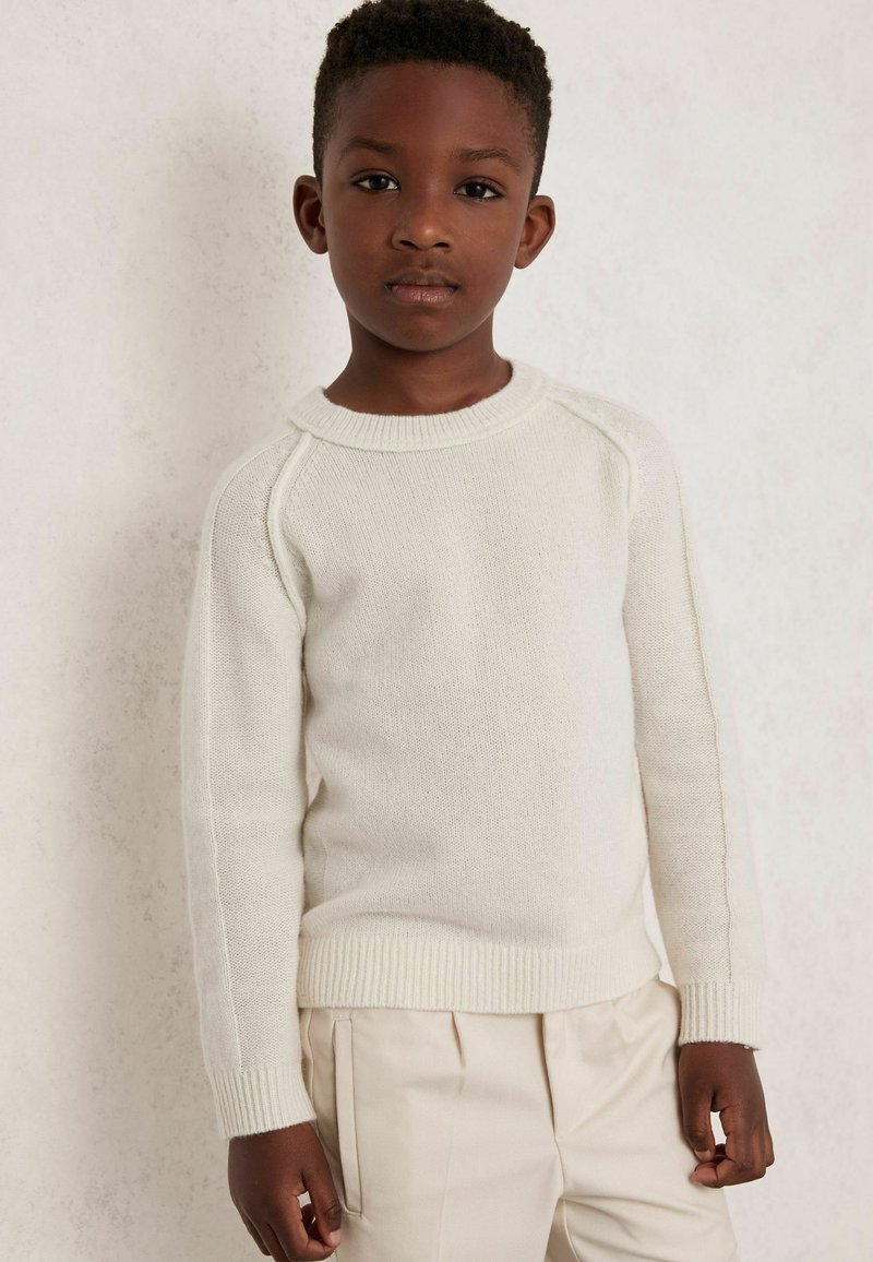 Svetlo bež pleteni pulover z okroglim izrezom, rebrastimi manšeti in robom, z raglan rokavi in mehko teksturo. Nosi se z svetlimi hlačami.