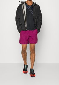 Veste imperméable noire avec des accents réfléchissants, chemise noire, shorts violets et chaussures noires avec des semelles oranges, bénéficiant d'un design respirant.