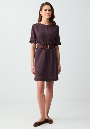 Donna che indossa un vestito viola a maniche corte con una larga cintura marrone e ballerine marroni, in piedi davanti a uno sfondo grigio chiaro uniforme.