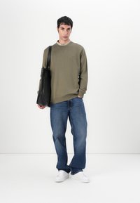 Olivengrønn genser med rund hals, kombinert med blå denimjeans. En svart totebag bæres i venstre hånd, supplert med hvite sneakers.