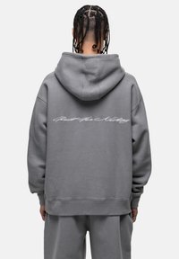 Felpa grigia con un grande cappuccio, vestibilità oversize, polsini e orlo a coste. Presenta testo bianco in corsivo sul retro. Tessuto morbido e resistente.