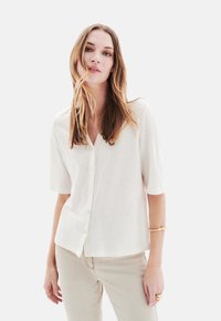 Helles beige, kurzärmeliges Button-Down-Oberteil mit V-Ausschnitt, aus einem weichen, strukturierten Stoff und in lässiger Passform. Kombiniert mit hellen Hosen.