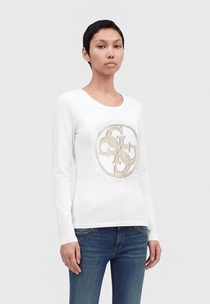 Femme portant un haut blanc à manches longues avec un emblème circulaire doré et un jean bleu, debout devant un fond gris clair uni.