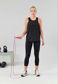 Crna majica bez rukava, crne tajice, drži crveni skakanje konopac, srebrne tenisice. Kettlebelli i bučice na svijetlozelenoj platformi.