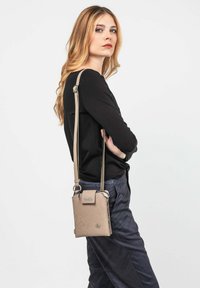 Junge Frau mit langen blonden Haaren, die ein schwarzes Langarmoberteil und navyblaue Hosen trägt. Sie hält eine beigefarbene Umhängetasche mit einem langen Schulterriemen.