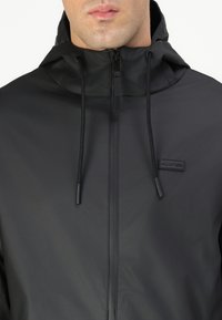 Black waterproof jacket with a zip closure, adjustable hood, and drawstrings. Features minimal branding and a sleek, smooth texture.