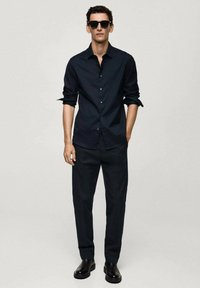 Camicia nera con bottoni e maniche arrotolate, abbinata a pantaloni scuri e stivali neri alla caviglia. Gli occhiali da sole completano l'outfit.