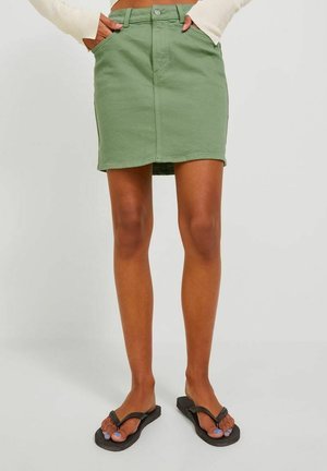 Femme portant une mini-jupe en denim verte, un pull crème et des tongs noires, debout les mains dans les poches de la jupe.