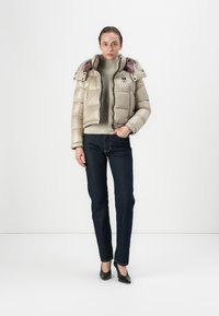 Beige pufferjack met een gestikt ontwerp, ritssluiting en een afneembare capuchon, gedragen over een lichte trui en donkere jeans. Zwarte hakken accentueren de outfit.