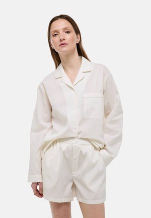 Femme portant une chemise blanche ample à boutons avec poche sur la poitrine et un short blanc assorti, debout devant un fond uni.