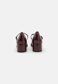 Talons en cuir verni bourgogne avec une forme arrondie, des brides ajustables et des talons carrés. Texture lisse avec un léger éclat.