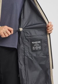 Texapore-jacket met een grijze binnenzijde en tan-afwerking. Voorzien van een ritszak met een label voor verzorgingsinstructies en een verstelbaar trekkoord.