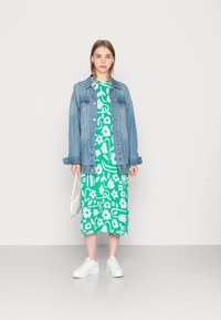 Jaqueta de ganga usada sobre um vestido verde com padrão floral. Ténis brancos e uma mala verde-clara completam o look. Texturas suaves e casuais.
