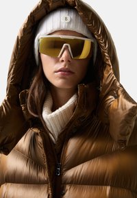 Kobieta w dużych lustrzanych goglach narciarskich, białej dzianinowej czapce, białym swetrze oraz błyszczącej brązowej kurtce puchowej z założonym kapturem.