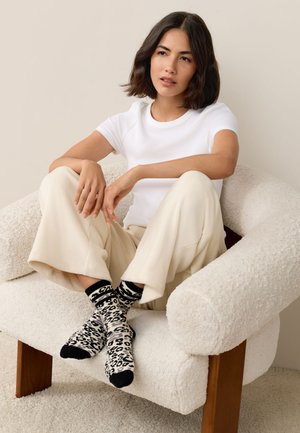 Woman with dark hair wearing a white t-shirt, cream wide-leg pants, and black-and-white leopard print socks, sitting cross-legged on a textured armchair.
