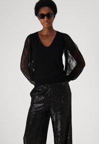 Black long-sleeve top with a V-neck and sheer textured sleeves paired with shiny black sequin trousers. Sunglasses complete the look.