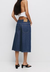 Les culottes en denim présentent une teinte bleu foncé, une taille mi-haute et deux poches arrière. Le tissu a une texture lisse, et un patch de marque en cuir marron est visible.