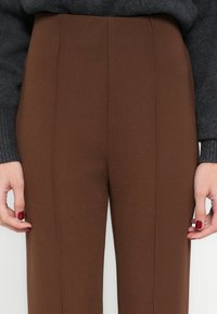 Hnedé na mieru šité nohavice s vysokým pásom, hladkou textúrou a prednými záhybmi. Tkanina je ľahká a nohavice majú rovný strih.