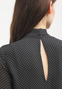Blusa nera a pois con collo alto, dettaglio cut-out sul retro e tessuto trasparente.