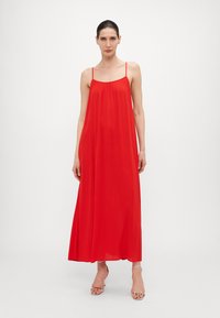 Červené maxi šaty s tenkými ramienkami, splývavou tkaninou a voľným strihom. Majú zberaný výstrih a hladký, jemný textil.