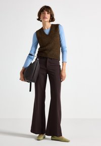 Gilet di maglia marrone sopra una maglietta a maniche lunghe azzurra, abbinato a pantaloni svasati marroni scuri e scarpe verdi. Tiene una borsa nera texturizzata.