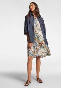 Džínová košile přehodnocená přes vzorované šaty v modrých, žlutých a zelených odstínech. Šaty mají volný siluetu s zakřiveným lemem. Hnědé sandály.