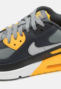 Nike Air Max 90 tossud, millel on hall ja must nahkpealne, kollased aktsendid ja hallid paelad. Õhupehmendus on nähtav tallas.
